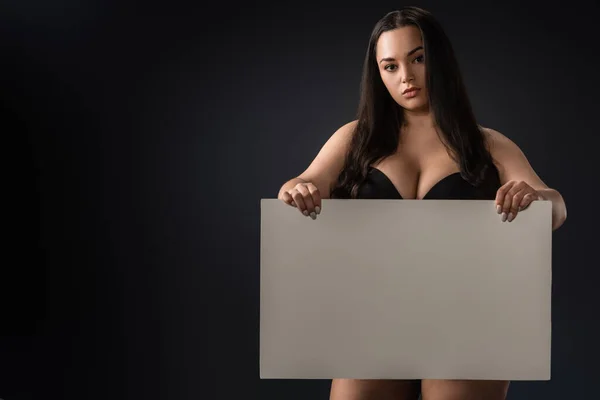 Beautiful plus size girl in bra with empty board on black background — Stock Photo