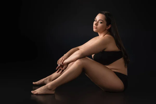 Vista laterale di bella plus size ragazza in biancheria intima seduta su sfondo nero — Foto stock