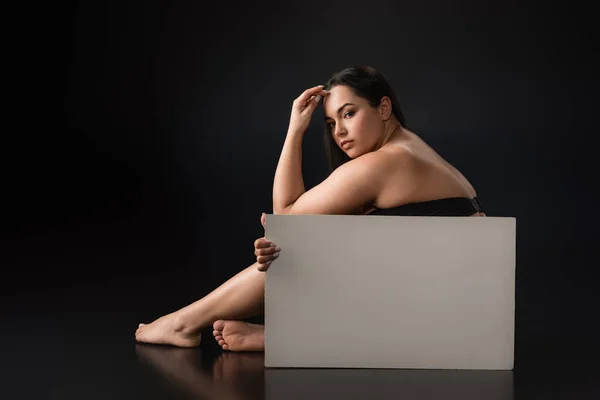 Vista frontal da menina bonita plus size sentado e segurando cartaz em branco no fundo preto — Fotografia de Stock