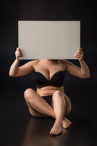 Front view of plus size model covering face with blank placard and sitting on black background — Stock Photo