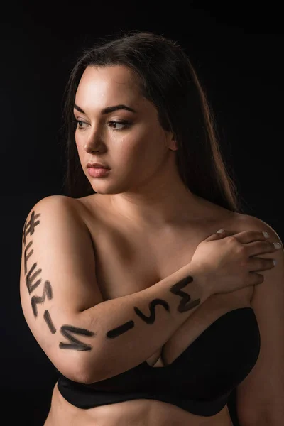 Retrato de modelo de talla grande en sujetador negro con etiqueta de letras feminismo en el cuerpo aislado en negro - foto de stock