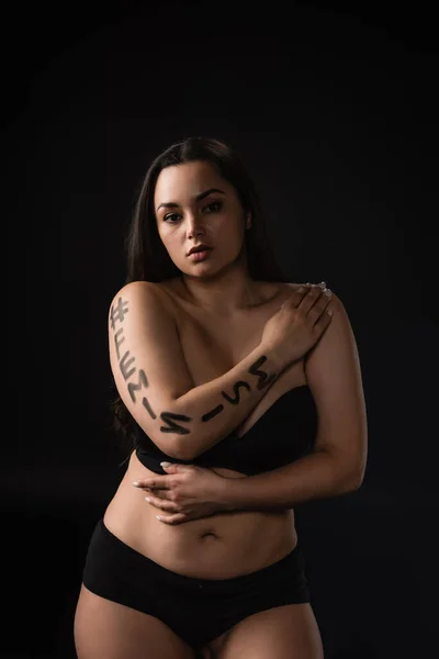 Front view of plus size model in black underware with lettering hashtag feminism on body isolated on black — Stock Photo