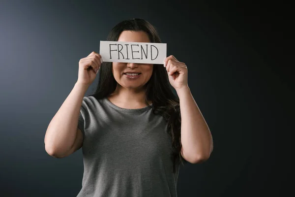 Вид спереди улыбающейся девушки plus size, закрывающей лицо карточкой со словом friend на чёрном фоне — стоковое фото