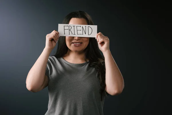 Vista frontal de la sonriente chica de tamaño grande cubriendo la cara con tarjeta con la palabra amigo sobre fondo negro — Stock Photo