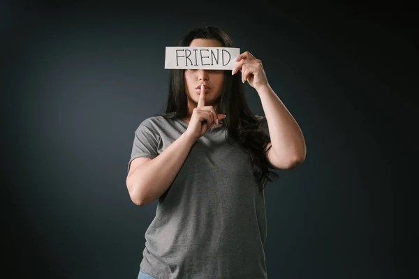 Front view of plus size girl covering face with card with word friend and silence gesture on black background — Stock Photo