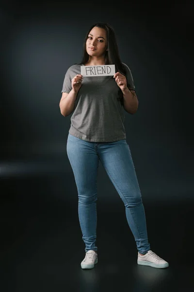 Front view of smiling plus size girl holding card with word friend on black background — Stock Photo