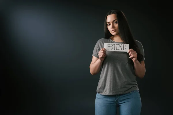 Front view of brunette plus size girl holding card with word friend on black background — Stock Photo