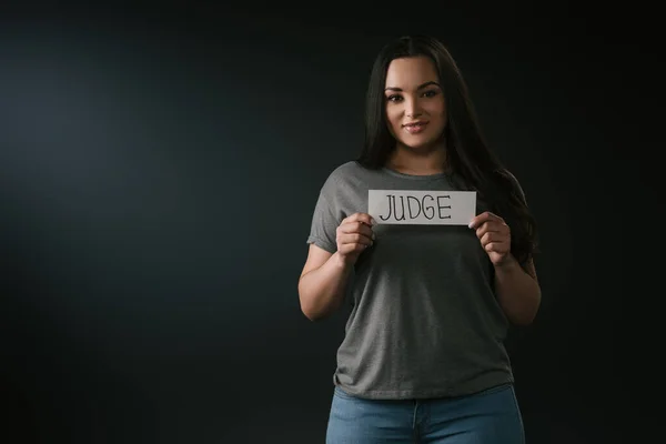 Вид спереди улыбающейся девушки plus size, держащей карточку со словом судья на черном фоне — стоковое фото