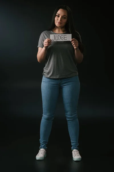 Full length view of smiling plus size girl holding card with word judge on black background — Stock Photo
