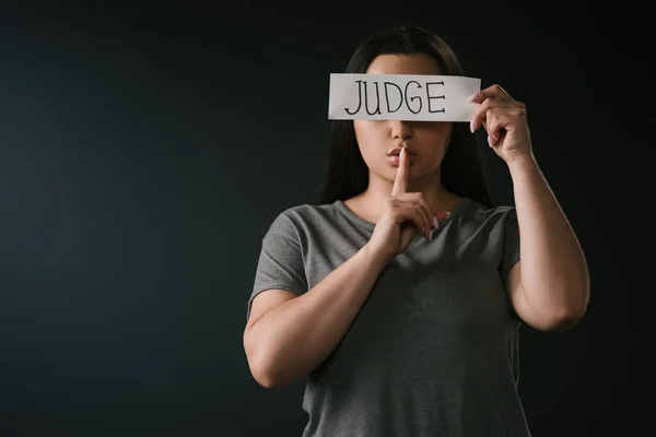 Front view of plus size girl covering eyes with card with word judge and silence gesture on black background — Stock Photo