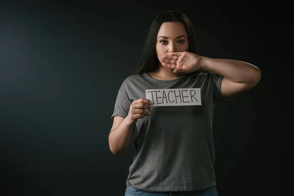 Vista frontale di plus size girl holding card con insegnante di parole e bocca di copertura con mano su sfondo nero — Foto stock