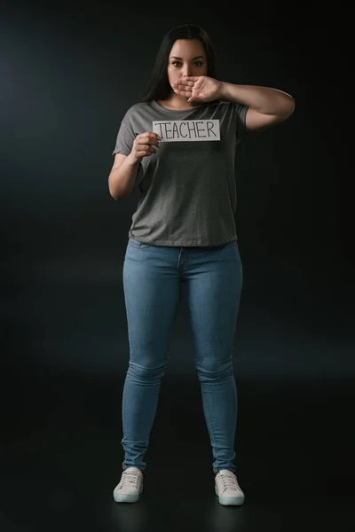 Visualizzazione full length di plus size girl holding card con insegnante di parole e bocca di copertura con mano su sfondo nero — Foto stock