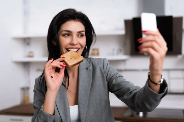Selective focus of smiling businesswoman with black dot on palm eating toast bread and taking selfie, domestic violence concept — Stock Photo