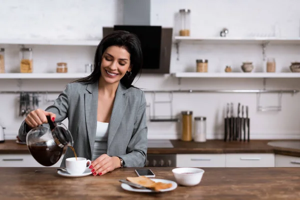 Smiling businesswoman pouring coffee in cup near smartphone with blank screen while hiding problem of domestic violence — Stock Photo