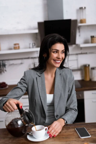 Donna d'affari sorridente versando caffè in tazza vicino allo smartphone con schermo bianco mentre nascondeva il problema della violenza domestica — Foto stock