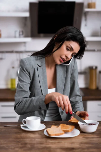 Красивая деловая женщина держит нож рядом с тостом хлеб, разговаривая на смартфоне и скрывая проблему домашнего насилия — стоковое фото