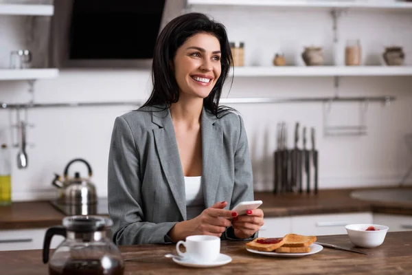 Cheerful businesswoman using smartphone near breakfast and hiding problem of domestic violence — Stock Photo