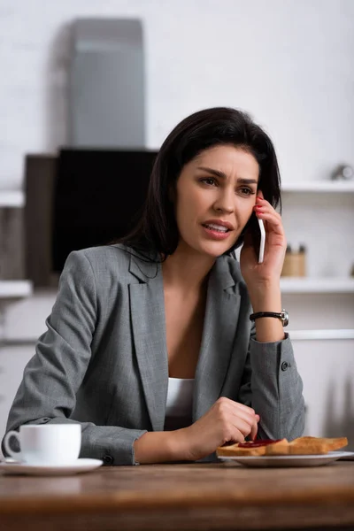 Enfoque selectivo de la mujer de negocios disgustado hablando en el teléfono inteligente cerca del desayuno - foto de stock