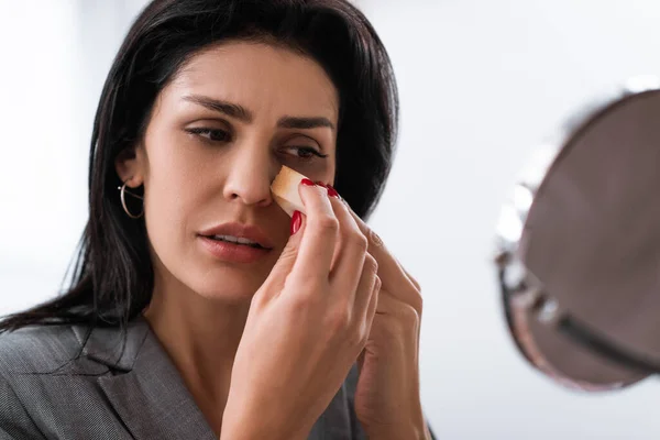 Selective focus of sad woman with bruise on face holding makeup sponge while applying makeup foundation near mirror, domestic violence concept — Stock Photo