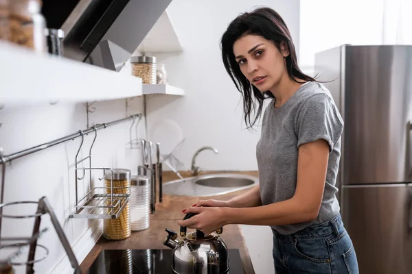 Selective focus of woman with bruise on face touching teapot on electric stove in kitchen, domestic violence concept — Stock Photo