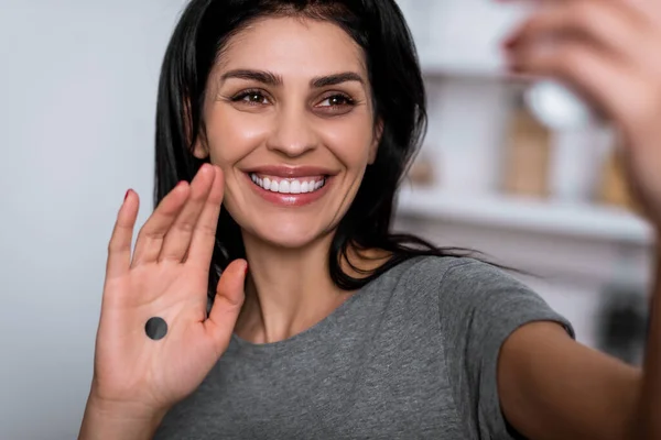 Selective focus of smiling woman with bruise on face and black dot on palm taking selfie at home, domestic violence concept — Stock Photo