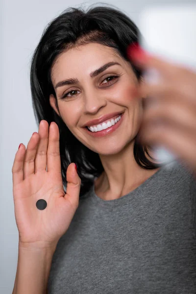 Selective focus of positive woman with bruise on face and black dot on palm taking selfie at home, domestic violence concept — Stock Photo