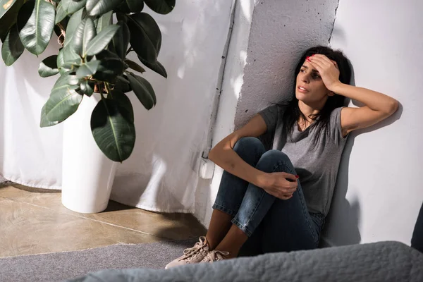 Enfoque selectivo de la mujer deprimida con moretones tocando la cara y sentado en el suelo cerca de la planta, concepto de violencia doméstica - foto de stock