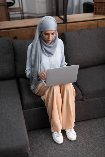 Mujer árabe en hijab usando computadora portátil en la sala de estar - foto de stock