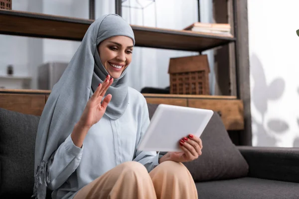 Mulher árabe positivo no hijab segurando tablet digital e mão acenando enquanto tem chamada de vídeo na sala de estar — Fotografia de Stock