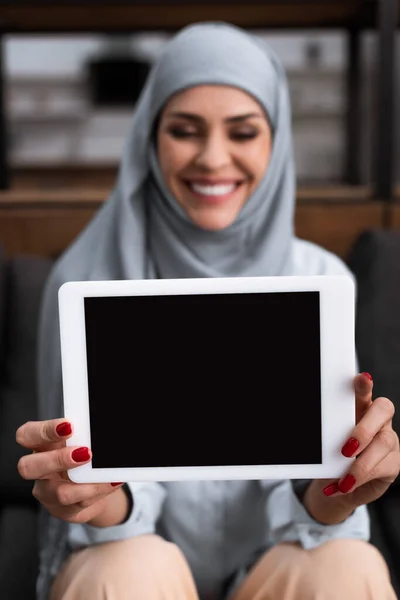 Enfoque selectivo de la mujer árabe positiva en hijab celebración tableta digital con pantalla en blanco en la sala de estar - foto de stock