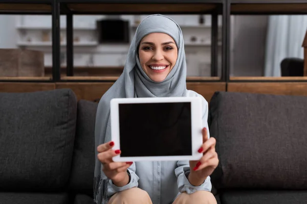 Selektiver Fokus einer lächelnden arabischen Frau im Hijab, die ein digitales Tablet mit leerem Bildschirm im Wohnzimmer hält — Stockfoto