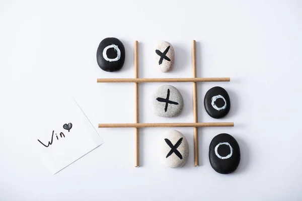 Top view of tic tac toe game with grid made of paper tubes, pebbles marked with crosses and naughts, and card with win inscription on white surface — Stock Photo
