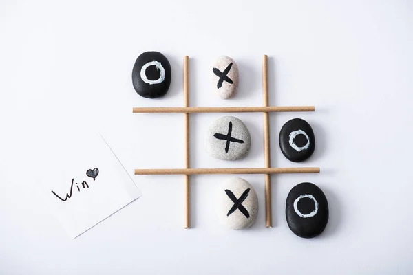 Vue du haut du jeu tic tac toe avec grille en tubes de papier, cailloux marqués de croix et de nausées, et carte avec inscription victoire sur surface blanche — Photo de stock