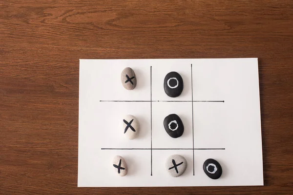 Верхний вид игры крестики-нолики на белой бумаге с камнями, отмеченными морскими и крестами на деревянной поверхности — стоковое фото