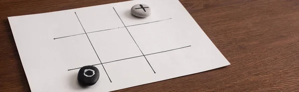 Panoramic shot of tic tac toe game on white paper with pebbles marked with naught and cross on wooden surface — Stock Photo