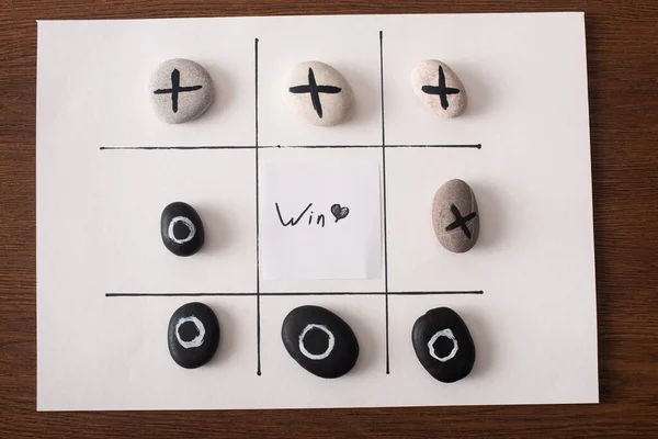 Top view of tic tac toe game on white paper with pebbles marked with naught and cross, and win inscription on wooden surface — Stock Photo