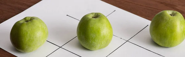 Shote panorámico de tic tac toe juego en papel blanco con fila de tres manzanas verdes en la superficie de madera - foto de stock