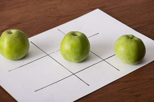 Крестики-нолики игры на белой бумаге с рядом из трех зеленых яблок на деревянной поверхности — стоковое фото