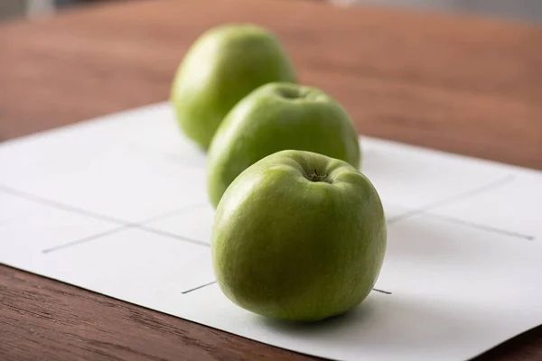 Enfoque selectivo de tic tac toe juego con fila de tres manzanas verdes sobre papel blanco en la superficie de madera - foto de stock