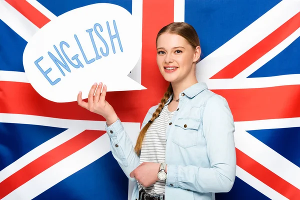 Smiling pretty girl with braid holding speech bubble with English lettering on uk flag background — Stock Photo
