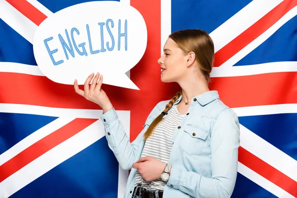 Sorrindo menina bonita com trança segurando bolha de fala com letras em inglês no fundo da bandeira do Reino Unido — Fotografia de Stock