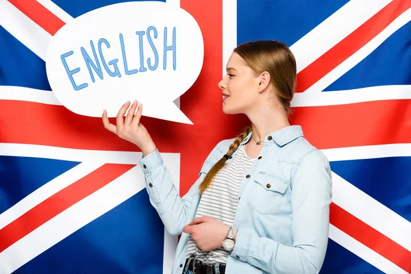 Smiling pretty girl with braid holding speech bubble with English lettering on uk flag background — Stock Photo