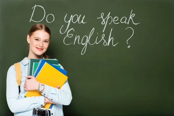 Smiling girl with copybooks and backpack standing near chalkboard with do you speak English lettering — Stock Photo