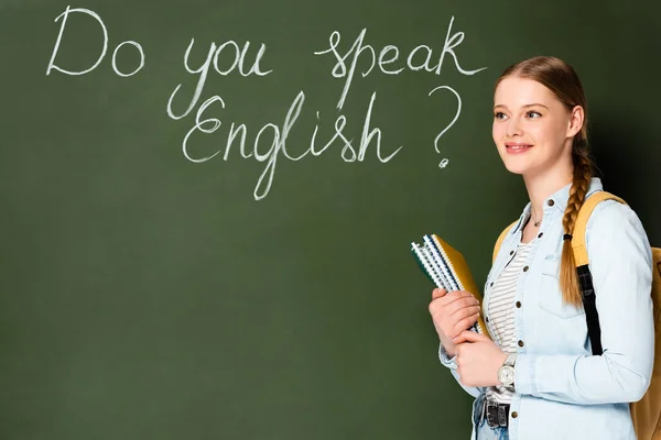 Smiling girl with copybooks and backpack standing near chalkboard with do you speak English lettering — Stock Photo