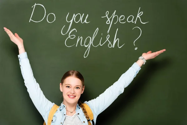 Smiling girl with backpack pointing at chalkboard with do you speak English lettering — Stock Photo