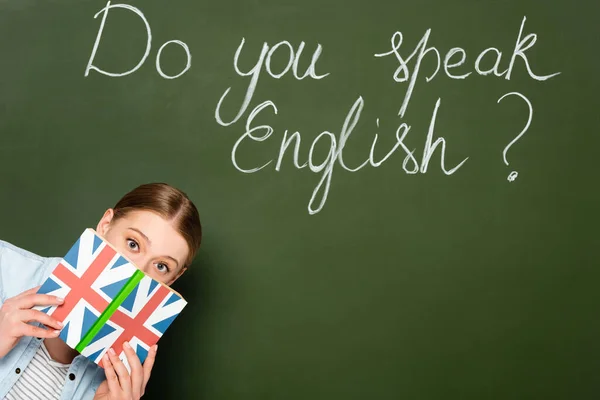 Hübsches Mädchen mit undurchsichtigem Gesicht, das Buch mit UK-Flagge in der Nähe der Kreidetafel hält, mit englischem Schriftzug — Stockfoto