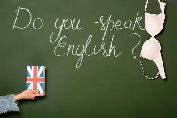 Cropped view of girl holding book with uk flag near chalkboard with do you speak English lettering and bra — Stock Photo