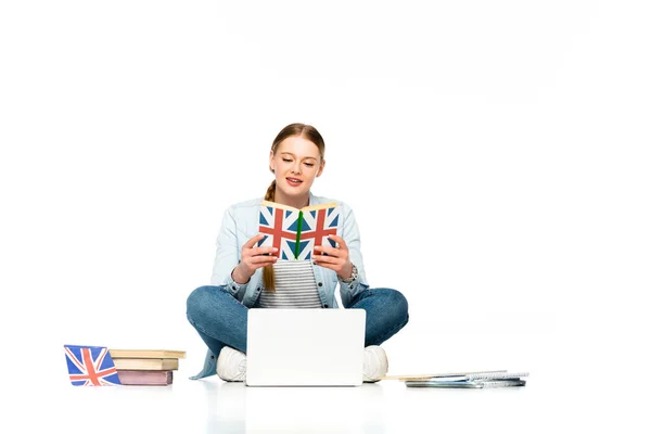 Menina sentada no chão com laptop, copybooks e bandeira do Reino Unido enquanto lendo livro isolado no branco — Fotografia de Stock