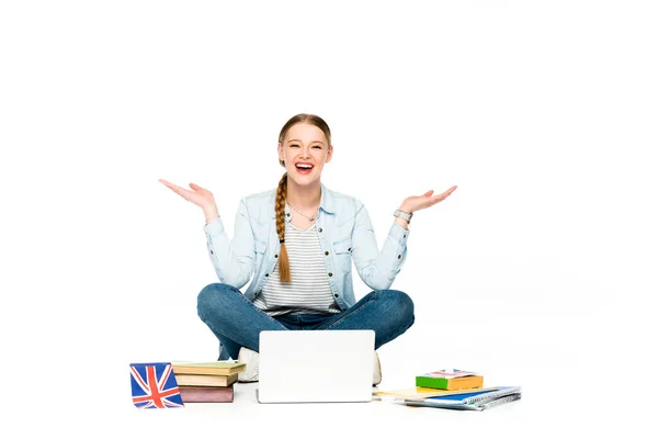 Glückliches Mädchen, das mit Laptop, Büchern und Textbüchern auf dem Boden sitzt, eine britische Flagge trägt und isoliert auf weiß gestikuliert — Stockfoto