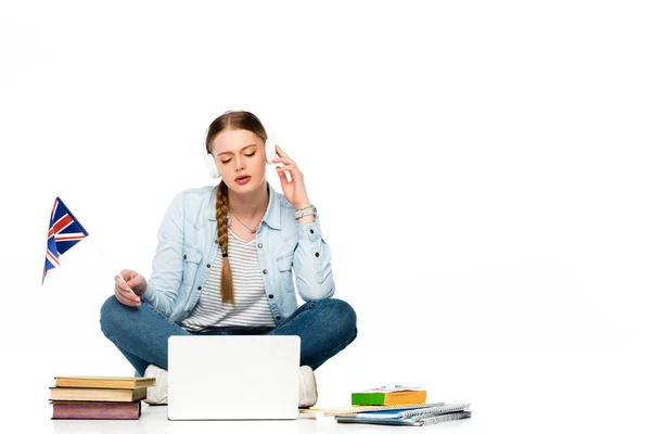 Niña sentada en el suelo en auriculares cerca de la computadora portátil, libros y copybooks, sosteniendo la bandera del Reino Unido aislado en blanco - foto de stock